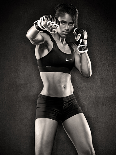 A black and white image of a woman in a sports bra and shorts, striking a boxing pose with one arm extended forward. She is wearing boxing gloves and has an intense, focused expression.