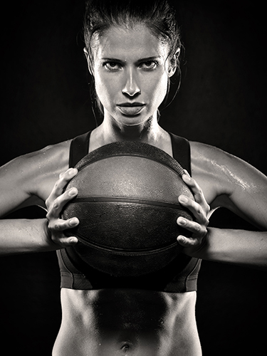 A woman holds a basketball with both hands. She is wearing a sports bra and appears focused and determined. Her skin glistens with sweat, indicating physical exertion.