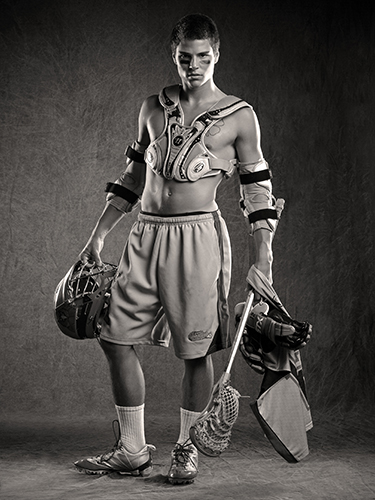 A black and white image of a young man in lacrosse gear. He holds a lacrosse stick and helmet, wearing shoulder and arm pads, shorts, and cleats.