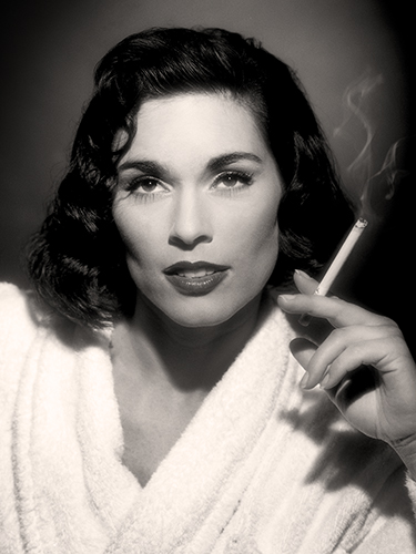 Portrait of a woman with wavy dark hair, wearing a white robe, holding a lit cigarette. The background is softly blurred, creating a classic vintage atmosphere.