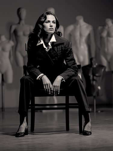 A woman in a dark suit sits confidently on a chair in a dimly lit room, surrounded by mannequins. The mood is sophisticated and stylish, highlighting fashion and elegance.