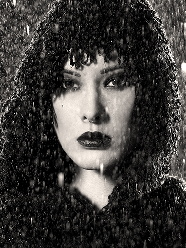 A woman with dark makeup and a textured head covering stares intensely forward. The image is in black and white, with rain falling heavily around her, creating a dramatic and moody atmosphere.