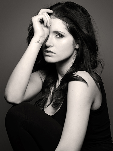 Black and white portrait of a woman with long dark hair, seated. She is wearing a sleeveless top and has her hand resting on her head, partially covering her eye. Her expression is serene as she looks towards the camera.