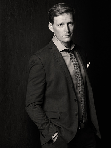 Black and white photo of a man in a suit and tie. He stands with one hand in his pocket, gazing at the camera. The background is dark, creating a dramatic contrast with his light hair and features.