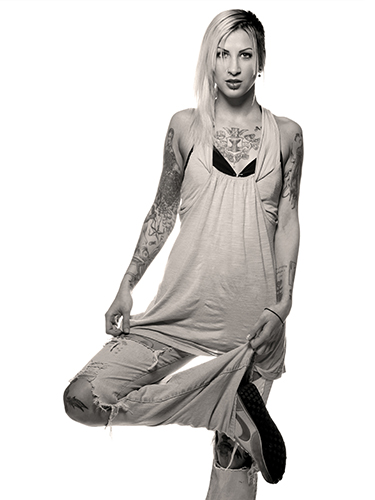 A woman with tattoos poses in a relaxed manner, wearing a loose tank top and ripped jeans. She is balances on one leg with the other folded, displaying intricate arm tattoos and a confident expression.