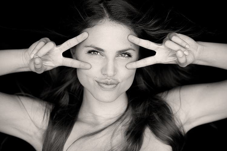 Black and white photo of a woman with long hair, playfully holding up two fingers on each hand in a peace sign near their eyes while puckering their lips.