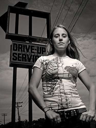 Portrait of a woman standing confidently in front of a vintage drive-up service sign. She wears a T-shirt with a skeletal and heart design. The sky is cloudy, and utility poles are visible in the background. 