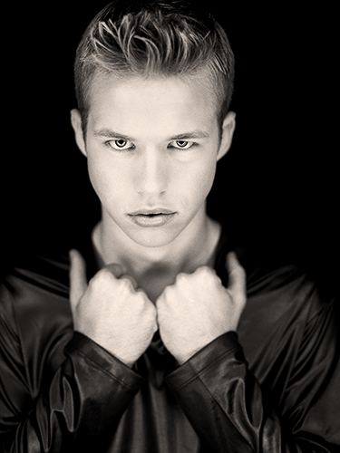 A man with short hair, wearing a dark, shiny jacket, looks directly into the camera with a serious expression. Their hands are raised to their chest, with fists slightly clenched, against a dark background.