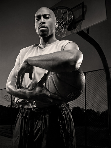 A man stands on an outdoor basketball court holding a basketball under one arm. He is wearing a sleeveless shirt and shorts. The background features a basketball hoop and a chain-link fence.