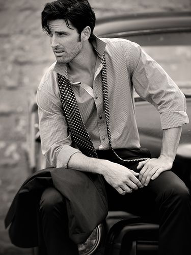 A man sits on the fender of a vintage truck, looking to the side. He wears a striped shirt with an open collar, tie loosely draped around his neck, and holds a jacket. The background is blurred, highlighting a rustic wall.