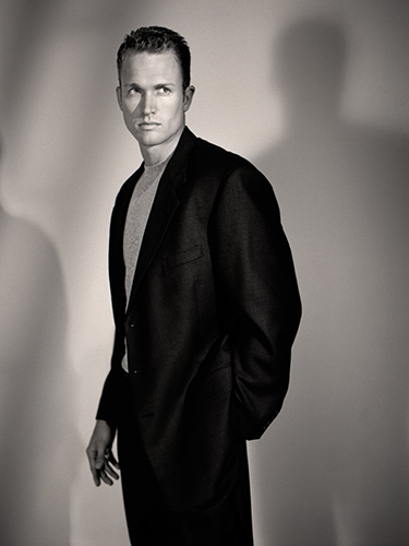 A man in a dark suit and light shirt stands confidently against a plain background. His hands are in his pockets, and his shadow is cast on the wall behind him. The image is in black and white, giving it a classic, timeless feel.