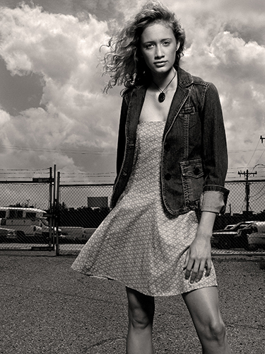 Black and white image of a woman standing outdoors on a cloudy day. She is wearing a denim jacket over a patterned dress. Her hair is blowing in the wind, and she stands on a paved area with a chain-link fence and vehicles in the background.