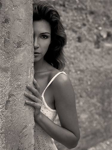 A woman in a sleeveless top partially hides behind a rough-textured wall. She looks directly at the camera with an intense expression. The image is in black and white, highlighting the contrast between her skin and the stone.