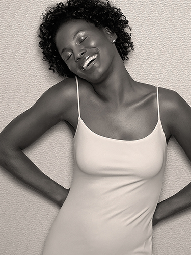 A woman with curly hair smiles joyfully, leaning back with her eyes closed. She is wearing a sleeveless dress and standing against a textured wall.