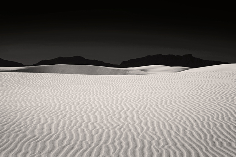 A desert landscape featuring vast sand dunes under a dark sky. The foreground shows textured ripples in the sand, while mountains are silhouetted in the background.