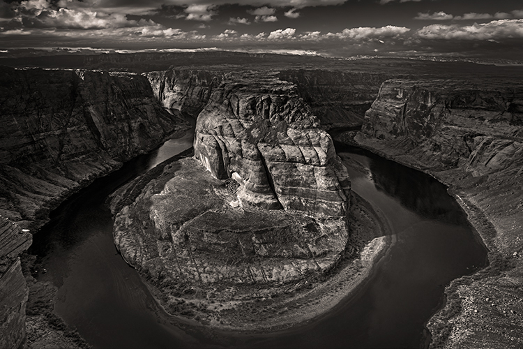 A dramatic black and white photo of Horseshoe Bend, where the Colorado River curves around a massive, rocky sandstone formation in a deep canyon under a cloudy sky.