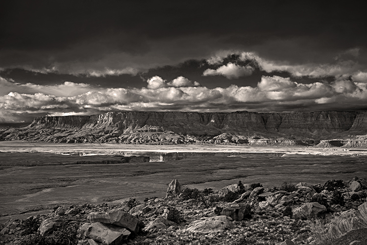 Black and white photo of a dramatic desert landscape with rocky terrain in the foreground, wide flat plains, distant cliffs, and a sky filled with thick, textured clouds.