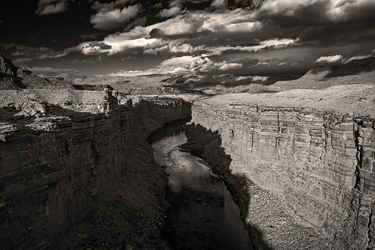 A dramatic black and white photo of a deep canyon with rugged rock walls and a winding river below, under a sky filled with clouds and sunlight breaking through.