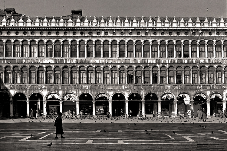 Black and white photo featuring the Procuratie facade of St Mark's Square in Venice. A few people and pigeons are scattered across the open plaza in front of the building. One person walks alone in the foreground.