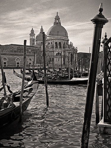Black and white photo of gondolas docked on a canal in Venice, with the domed Basilica di Santa Maria della Salute visible in the background and several wooden poles in the water.