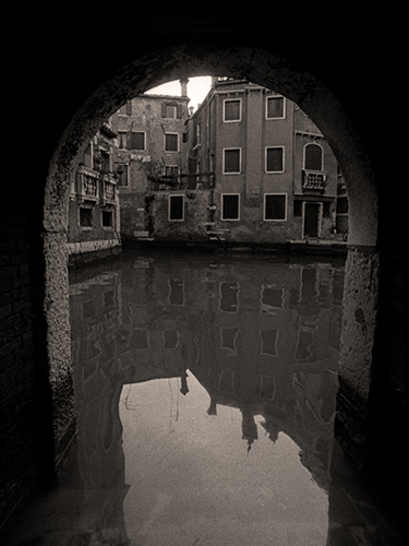 A stone arch frames a quiet canal in Venice, Italy. Old buildings with shuttered windows reflect on the still water, creating a peaceful, moody atmosphere in black and white.