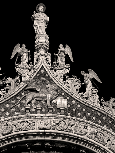 Ornate architectural detail atop of Saint Mark’s Basilica featuring a statue of St. Mark holding a book at the top, flanked by angels, with a winged lion below and intricate carvings and star patterns on the structure.