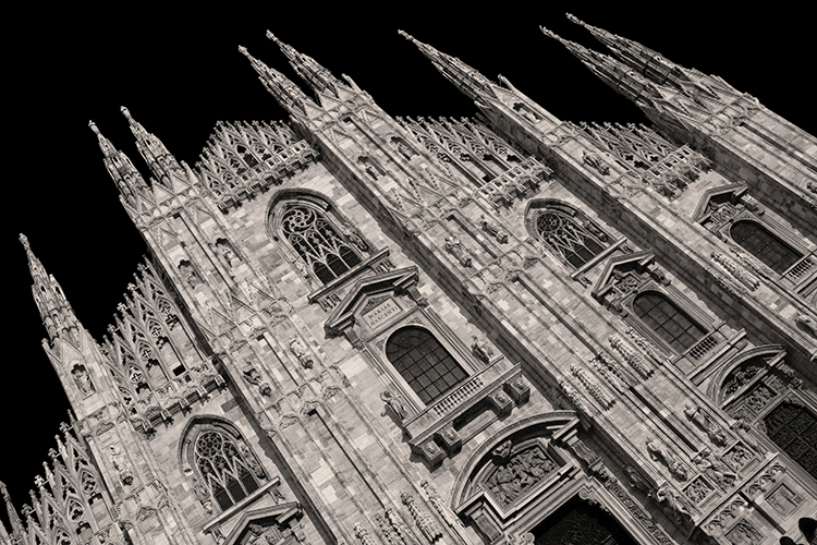 Black-and-white photo of the façade of Milan’s Gothic cathedral, featuring pointed arches, ornate stone carvings, spires, and large stained-glass windows against a solid black background.