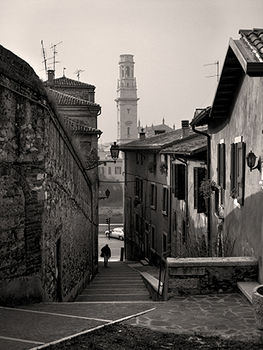 Black and white photo of a narrow, cobblestone street lined with old buildings. A lone figure walks down the stairs towards a distant bell tower. The scene appears to be in a historic European town.