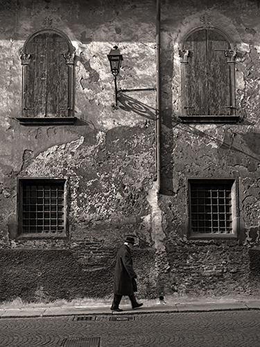 A person in a long coat and hat walks along a cobblestone street in front of a weathered building with two shuttered windows and two barred windows. A vintage street lamp casts a shadow on the textured wall.