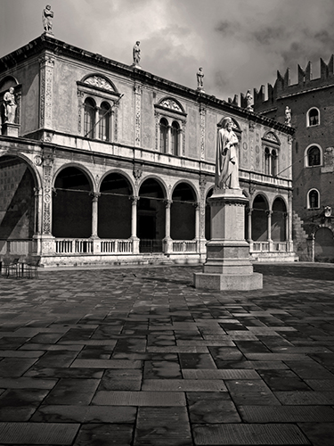 Black and white photo of an empty stone courtyard with a statue at its center, surrounded by a historic building featuring arches, columns, and several rooftop statues under a cloudy sky. 