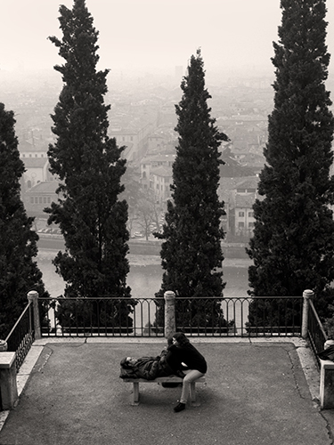 A couple sits on a bench, in a fenced overlook with tall trees and a hazy cityscape in the background. The scene is in black and white.