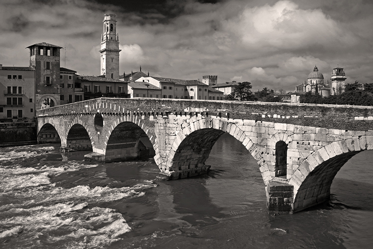 A black-and-white photo of a historic stone arch bridge spanning a river, with old buildings and a tall bell tower in the background under a cloudy sky.